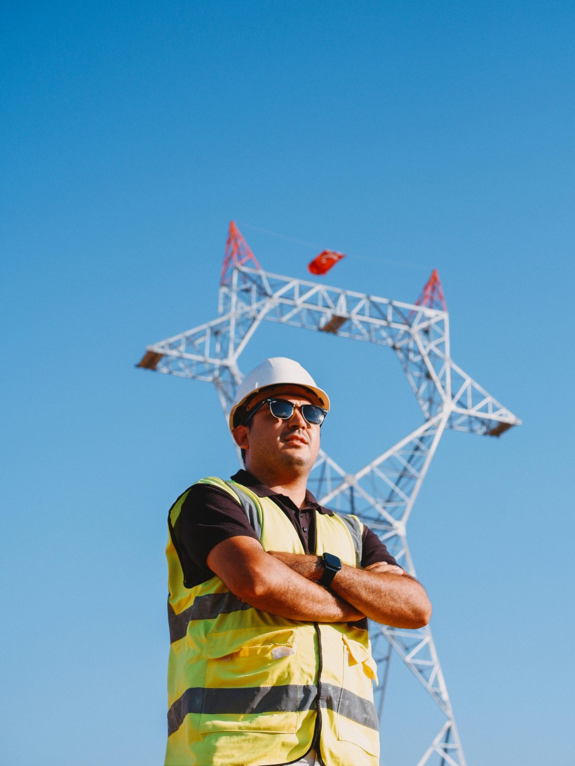 Confident construction worker in safety gear stands before a power line tower against a blue sky.