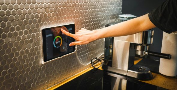 Modern kitchen scene featuring a hand using a tablet to control a smart home system.